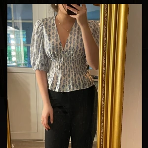 Vit blus med blått mönster och puffärm - Säljer en vit blus från H&M med blått blommigt mönster, puffiga korta ärmar och smockad midja. Blusen är aldrig använd med prislappen på!