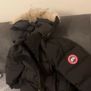 Svart dunjacka från Canada Goose - Säljer en svart dunjacka från Canada Goose med ikonisk patch på ärmen och stor huva med äkta pälskant. Jackan har dragkedja och tryckknappar framtill, samt flera praktiska fickor. Perfekt för kalla vinterdagar och riktigt snygg streetstil.