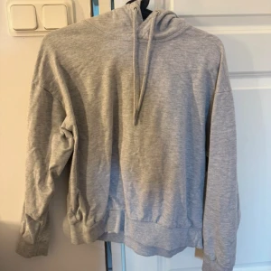 Grå hoodie med huva och snörning - En basic grå hoodie med huva och snörning. Tröjan har långa ärmar och ribbade muddar vid ärmslut och nederkant. Perfekt för chill dagar eller när du vill ha något bekvämt och stilrent.
