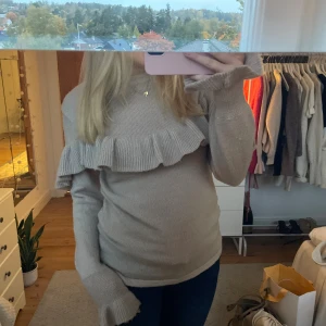 Beige stickad volangtröja - Superfin beige stickad tröja med volangdetalj över bröstet och axlarna. Hög hals och långa ärmar med extra volang vid ärmslutet. Perfekt för dig som gillar en söt och trendig look till hösten eller vintern.
