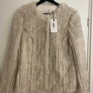 Beige pälsjacka (fusk) från Mayla - Snygg beige pälsjacka (fusk) från Mayla med två diskreta fickor i sidan samt knäppning (dolda)  framtill. Jackan har en fluffig, lyxig känsla och är helfodrad med slät insida. Storlek 38. Oanvänd, med prislapp kvar (inköpt på NK). Nypris 4250 kr. Perfekt för dig som vill sticka ut med en trendig och mjuk look. 