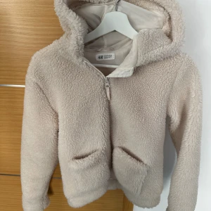 Beige teddyjacka med huva från H&M - Mysig beige teddyjacka från H&M med huva och dragkedja framtill. Jackan har två fickor och är tillverkad i ett fluffigt teddy-material som håller dig varm och skön. Perfekt för kyliga dagar när du vill ha en chill och trendig look.
