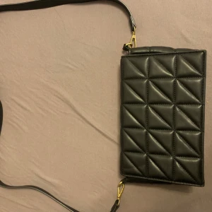 Svart quiltad axelväska - Snygg svart axelväska med quiltad design och guldfärgade detaljer. Väskan har en rektangulär form och justerbar axelrem. Perfekt för dig som vill ha en stilren och trendig accessoar till din outfit.