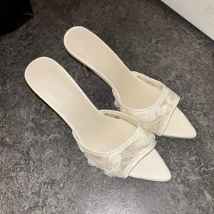 Vita spetsiga klackskor med spets - Säljer ett par vita pumps med spetsig tå och hög klack. Skorna har en öppen häl och är dekorerade med vacker spets och små blommor. Perfekta för dig som vill ha en elegant och romantisk look. Klacken är smal och ger en snygg siluett.