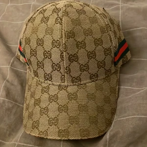Beige keps med GG-mönster och ränder - Snygg beige keps med all-over GG-logomönster och klassiska gröna och röda ränder på sidorna. Justerbar rem baktill med metallspänne. Kepsen är tillverkad i ett slitstarkt tyg och har en böjd skärm. Perfekt accessoar för en trendig streetstyle.