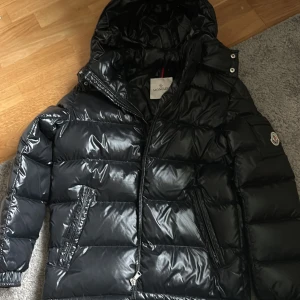 Moncler Maya barnjacka - Säljer en svart, glansig pufferjacka från Moncler med huva och dragkedja. Jackan har quiltad design, två snedställda fickor med dragkedja och klassisk Moncler-logga på ärmen. Perfekt för kalla dagar och riktigt snygg streetstil. Storleken är 14y. Aldrig använd.
