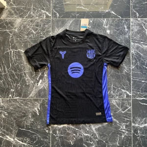 Svart Barcelona fotbollströja Nike - Snygg svart Barcelona fotbollströja från Nike med lila detaljer och klubbmärke på bröstet. Tröjan har korta ärmar, rund hals och är tillverkad i ett lätt, ventilerande funktionsmaterial. Spotify-logga framtill och diskret mönster i tyget.