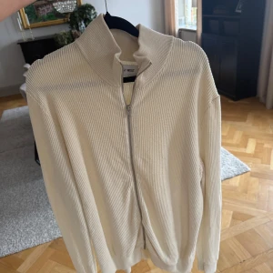 Beige ribbad kofta från East West - Ribbstickad beige kofta från East West med hög krage och hel dragkedja framtill. Långärmad och i mjukt material som passar perfekt till lager-på-lager. Enkel och clean design som funkar till allt.