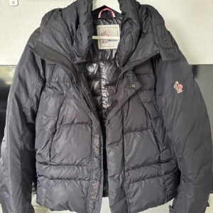  Moncler pufferjacka med huva - Svart pufferjacka från Moncler med stor, mysig huva och broderad logga på ärmen. Jackan har quiltad design, dragkedja och knappar framtill. Materialet är glansig polyester som ger en clean och modern vibe. Perfekt för kalla dagar när du vill hålla stilen. Den är köp på kasperheat för ett år sendan använd den väldigt lite för de passar inte.