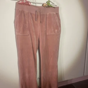 Rosa velourbyxor Juicy Couture M - Supermjuka rosa velourbyxor från Juicy Couture med bred resår och snörning i midjan. Byxorna har stora fickor fram och bak, samt broderad JC-logga på ena fickan. Perfekta för en chill och trendig look.