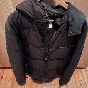 Svart pufferjacka från Antony Morato - Svart pufferjacka från Antony Morato med huva och dragkedja framtill. Jackan har quiltad design och logotypband i svart och gult över bröstet och ryggen. Perfekt för kalla dagar och har en modern, sportig vibe.