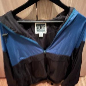 Blå och svart vindjacka från Colors - Snygg vindjacka från Colors i blått och svart med dragkedja framtill och elastiska muddar. Jackan har en luva och är tillverkad i lätt polyester, perfekt för blåsiga dagar. Klassisk loose passform och sportig look. Fläck på ena ärmen 