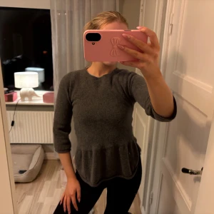 Grå stickad topp från By Malene Birger - Mysig grå stickad topp från By Malene Birger med peplumvolang nedtill och trekvartsärm. Enkel och stilren design med rund halsringning, perfekt att matcha med jeans eller kjol. Materialet är mjukt och skönt mot huden.