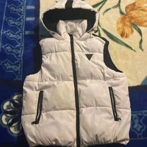 Vit dunväst med huva från Guess XS - Snygg vit dunväst från Guess i storlek XS med svart detaljer och huva. Västen har två fickor med dragkedja, svart Guess-logga på bröstet och quiltad design. Perfekt för lager-på-lager och streetstyle. Stängs med dragkedja framtill.