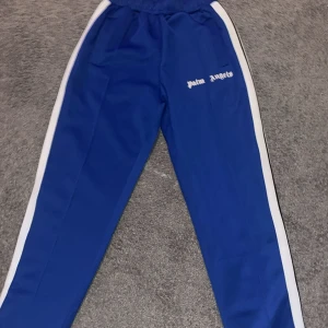 Palm angels  - Snygga blå trackpants från Palm Angels med vita sidoränder och logga broderad på vänster ben. Byxorna har elastisk midja och raka ben, tillverkade i glansig polyester för en sportig vibe. Perfekta för en streetwear-look.