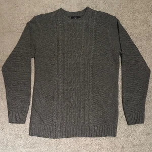 Mörkgrå stickad tröja med kabelmönster - Snygg mörkgrå stickad tröja med klassiskt kabelmönster framtill och ribbade muddar vid ärmslut och nederkant. Tröjan har rund hals och långärmade ärmar, perfekt för dig som gillar en enkel men stilren look.