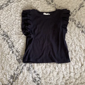 Svart topp med volangärmar - Svart topp med rund halsringning och snygga volangdetaljer på axlarna. T-shirten är ärmlös och har en enkel, stilren design som passar till många olika outfits. Perfekt för dig som gillar en trendig och feminin look.