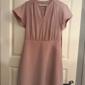 Ljusrosa klänning från H&M, strl 40 - Säljer en ljusrosa klänning från H&M i storlek 40. Klänningen har korta ärmar, v-ringad hals och markerad midja med lätt rynkning. Materialet är mjukt och slätt, troligen polyester. Dragkedja i sidan för enkel påklädning. Enkel och stilren design. Använd fåtal gånger. Superfin klänning med fina detaljer. Den är tillsydd i urringningen men är lätt och enkelt att spreta upp. 