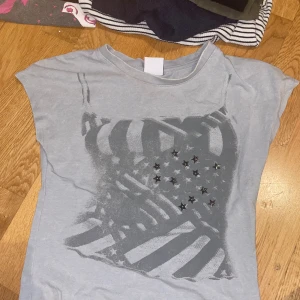 Ljusgrå t-shirt med stjärndetaljer - Snygg ljusgrå t-shirt med korta ärmar och tryck av amerikansk flagga i grå toner. Framsidan är dekorerad med små svarta stjärnor som ger en cool touch. Perfekt för dig som gillar streetstyle och unika detaljer.