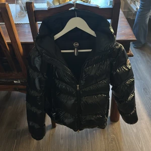 Svart pufferjacka från Colmar - Säljer en svart pufferjacka från Colmar med glansig finish och stor huva. Jackan har dragkedja framtill, två sidofickor med dragkedja och en schysst puffig look. Perfekt för dig som gillar streetstyle och vill hålla dig varm under kalla dagar.