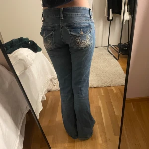 Blå bootcut jeans med broderade fickor - Säljer ett par blå bootcut jeans med låg midja och snygga broderade detaljer på bakfickorna. Jeansen har dubbla knappar framtill och klassiska fickor både fram och bak. Perfekta för dig som gillar en retro vibe och vill sticka ut lite extra. 