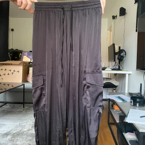 Svarta cargopants i satin-look - Snygga svarta cargopants med glansig satin-look och elastisk midja med snörning. Byxorna har stora fickor på sidorna och en avslappnad passform som ger en chill vibe. Perfekta för dig som gillar streetstyle och vill ha något bekvämt men ändå trendigt.