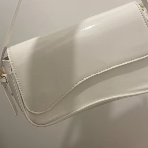 Vit axelväska i syntetmaterial - Snygg vit axelväska med stilren, böjd form och guldfärgade detaljer vid remmarna. Väskan har en slät, glansig yta och är tillverkad i syntetmaterial. Perfekt storlek för det viktigaste och enkel att matcha med olika outfits.