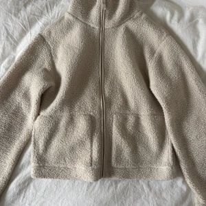 Beige teddyjacka med dragkedja XS - Mysig beige teddyjacka i storlek XS med hel dragkedja framtill och två stora fickor. Jackan har en avslappnad passform och är tillverkad i mjukt fleece-material som håller dig varm under kyliga dagar. Perfekt för lager-på-lager och chill vardagsstil.