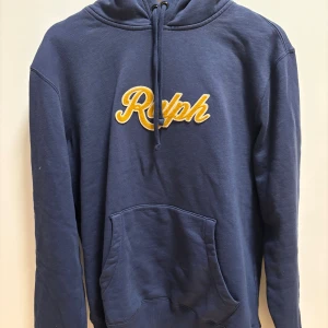 Polo Ralph Lauren Hoodie - Snygg marinblå hoodie från Polo Ralph Lauren med broderad gul text 'Ralph' på bröstet. Tröjan har huva med snörning och en stor magficka. Perfekt för dig som gillar klassisk streetstyle och vill ha något bekvämt och stilrent. Nypris 3200kr