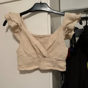 Beige omlott linnetopp med volang strl: XS - Säljer en beige linnetopp med omlott-design framtill och volangdetaljer på axelbanden. Toppen är croppad och har en lätt rynkad struktur i materialet som ger en chill och somrig vibe. Perfekt för dig som gillar ljusa färger och mjuka material.
