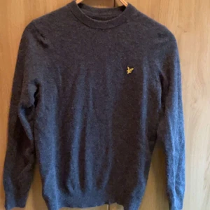 Grå stickad tröja från Lyle & Scott - Mörkgrå stickad tröja från Lyle & Scott med rund halsringning och klassisk gul logga på bröstet. Tröjan har lång ärm och är perfekt för lager-på-lager under kyliga dagar. Enkel och stilren design som funkar till det mesta. Använd 1 gång 