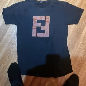 Svart Fendi t-shirt med logga - Svart t-shirt från Fendi med klassisk rund hals och korta ärmar. På bröstet sitter en stor brun Fendi-logga i skinnimitation som ger en lyxig vibe. Perfekt för dig som vill sticka ut med en ikonisk streetstyle-look. Samma tröja som duke dennis hade i rizalisa
