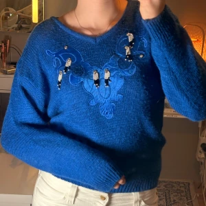 Blå stickad tröja med pärldetaljer - Säljer en blå stickad tröja från Spader Dam i storlek S. Tröjan har ett broderat mönster framtill och coola pärldetaljer som hänger ner från bröstet. Den har lång ärm, rund hals och ribbade muddar. Perfekt för dig som vill sticka ut lite extra! Några paljetter saknas men ingenting som är märkbart 