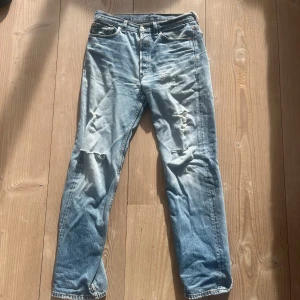  Levis 501 med slitningar - Ljusblå Levis 501 jeans med klassisk rak passform och snygga slitningar på knäna. Jeansen har fem fickor, knappgylf och är tillverkade i robust denim. Perfekta för en avslappnad stil. Storlek 32:32