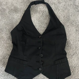 Svart halterneckväst - Svart väst med halterneck och djup v-ringning. Västen har fem knappar framtill och två dekorativa fickor. Passformen är slim och ryggen är öppen. Sitter som en S.