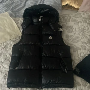 Svart dunväst  - Snygg svart dunväst  med klassisk logga på bröstet och dragkedja framtill. Västen har en hög krage och är perfekt för lager-på-lager-stil. Passar dig som vill ha en stilren och exklusiv look.