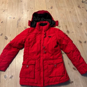 Röd dunjacka från Helly Hansen - Röd dunjacka från Helly Hansen med avtagbar huva, stora fickor med knappar och hög krage. Jackan har quiltad design och stängs med både dragkedja och knappar. Perfekt för kalla dagar och snygg streetstil.