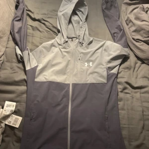 Grå vindjacka från Under Armour M - Snygg tvåfärgad vindjacka från Under Armour i ljusgrått och mörkgrått. Jackan har huva, hel dragkedja, bröstficka med dragkedja och logga på bröstet. Elastiska muddar med broderad text på ärmen. Perfekt för aktiva dagar.