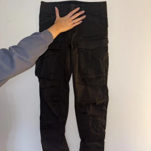 Svarta byxor från G-Star Raw - Svarta byxor från G-Star Raw med flera stora fickor på benen och dragkedjedetaljer. Byxorna har tapered passform och är tillverkade i bomull med lite stretch. Perfekta för en streetwear-look och riktigt sköna att bära hela dagen.