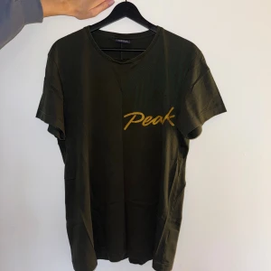 Mörkgrön Peak Performance t-shirt - Snygg mörkgrön t-shirt från Peak Performance med gul text 'Peak' på bröstet. Modellen har korta ärmar, rund hals och är tillverkad i 100% bomull. Diskret mönster i tyget ger en cool touch. Perfekt för dig som gillar streetwear och sportiga vibes.