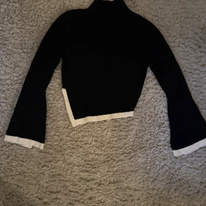 Svart polotröja med vita detaljer - Snygg svart polotröja med vita kanter på ärmslut och nederkant. Tröjan är ribbstickad, har lång ärm och en croppad, asymmetrisk passform som ger en cool och modern look. Perfekt för dig som gillar stilrena plagg med en twist.