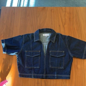 Mörkblå croppad jeansjacka från Zara - Säljer en mörkblå croppad jeansjacka från Zara med korta ärmar och två stora fickor framtill. Jackan har klassisk krage, kontrastsömmar och en loose passform. Perfekt för dig som gillar streetstyle och vill ha en chill look.
