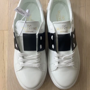 Valentino Garavani Open sneakers vit/blå - Snygga Valentino Garavani Open sneakers i vitt skinn med bred marinblå läderdetalj över snörningen och ikoniska nitar baktill. Klassisk rund tå, platt sula och vita skosnören. Guldiga logotyper på plösen och innersulan. Perfekta för dig som gillar lyxiga och stilrena sneakers.