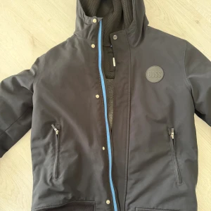 Svart BARN vinterjacka från Hugo Boss - Svart vinterjacka från Hugo Boss med huva, blå dragkedja och två fickor med dragkedja. Jackan har ett stort BOSS-märke på bröstet och meshfoder på insidan. Perfekt för dig som vill ha en stilren och sportig look. Barnstorlek 14år