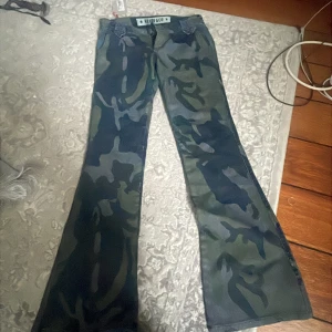 Camo bootcut byxor från Staff & Co S - Snygga camouflage-mönstrade byxor från Staff & Co i bootcut-modell. Byxorna har stora fickor bak med lock och knapp, samt coola detaljer vid hällorna. Färgerna går i grönt, blått och svart. Perfekta för dig som vill sticka ut med en edgy stil.