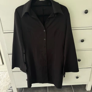 Svart långärmad skjortklänning  - Stilren svart skjortklänning med lång ärm och klassisk krage. Skjortklänningen har vida ärmar och knäppning hela vägen framtill. Perfekt för dig som gillar minimalistisk och clean stil. Materialet känns mjukt och följsamt mot kroppen.