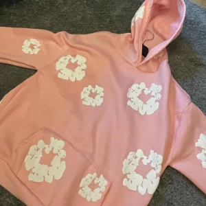 Rosa hoodie från Denim Tears med blommor - Säljer en rosa hoodie från Denim Tears med vita blommiga tryck över hela tröjan. Hoodien har en stor ficka framtill och huva. Perfekt för dig som vill sticka ut med en unik och färgglad stil.passar L eller en större M skulle jag säga Obs vet ej om den är ä.kta