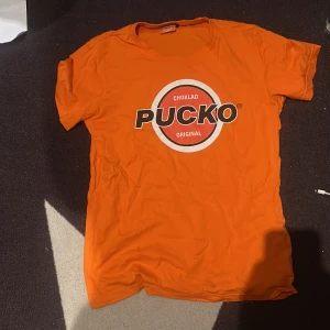 Orange Pucko t-shirt med tryck - Orange t-shirt från Pucko med stort tryck på bröstet där det står 'Choklad Pucko Original' i vitt, svart och rött. Klassisk rund hals och korta ärmar. Perfekt för dig som gillar retro och vill sticka ut med en färgstark look.