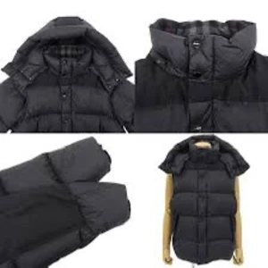 Svart dunväst med hög krage - Snygg svart dunväst med hög krage och avtagbar huva. Västen har breda, quiltade paneler och knäpps med både dragkedja och knappar framtill. Perfekt för lager-på-lager och håller dig varm under kyliga dagar.