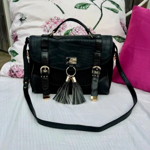 Svart axelväska från River Island - Snygg svart axelväska från River Island i nyskick. Väskan har ett krokodilpräglat mönster med  fransar och gulddetaljer. Delar av väskan i tyg. Den har både handtag och en justerbar axelrem. Ca 23x30 cm. Hämtas på Hägerstensåsen eller ök.